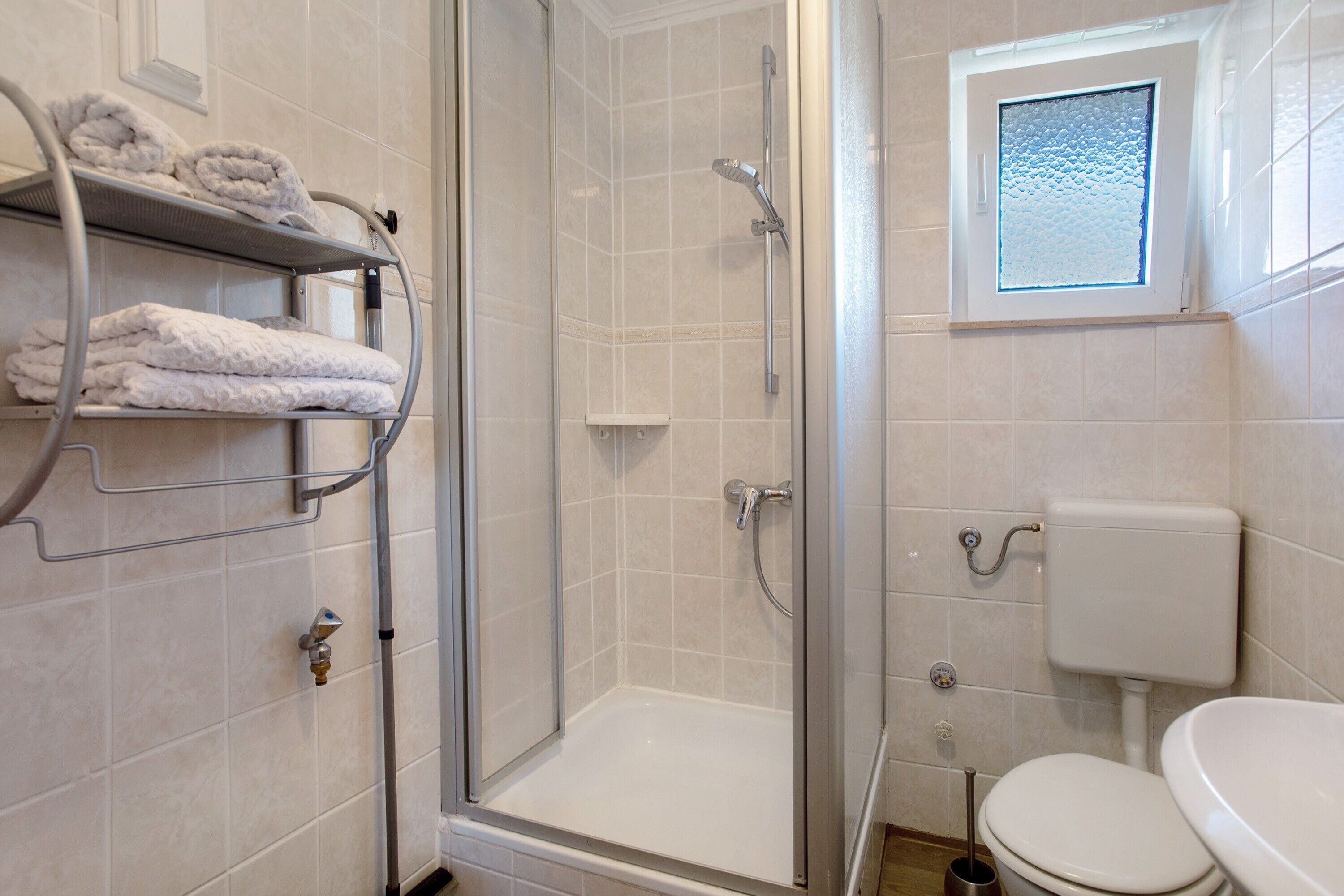 A bathroom with a glass-enclosed shower, a toilet, and a shelf with folded towels.