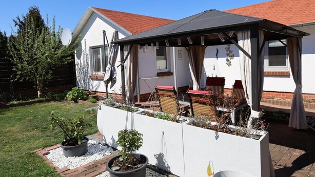 Een overdekt terras met tuinmeubilair, een witte muur met plantenbakken en een gebouw met een rood dak.