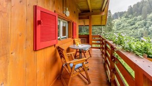A wooden balcony with red shutters, a table with chairs, and a view of a forested hillside.