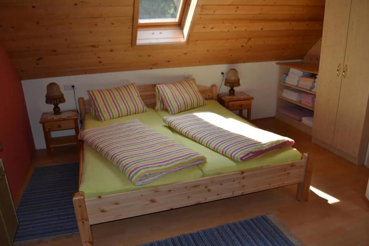 Een houten bed met twee kussens en een gestreept dekbed, een nachtkastje met een lamp, een klein plankje met opgevouwen handdoeken, en een houten kledingkast.
