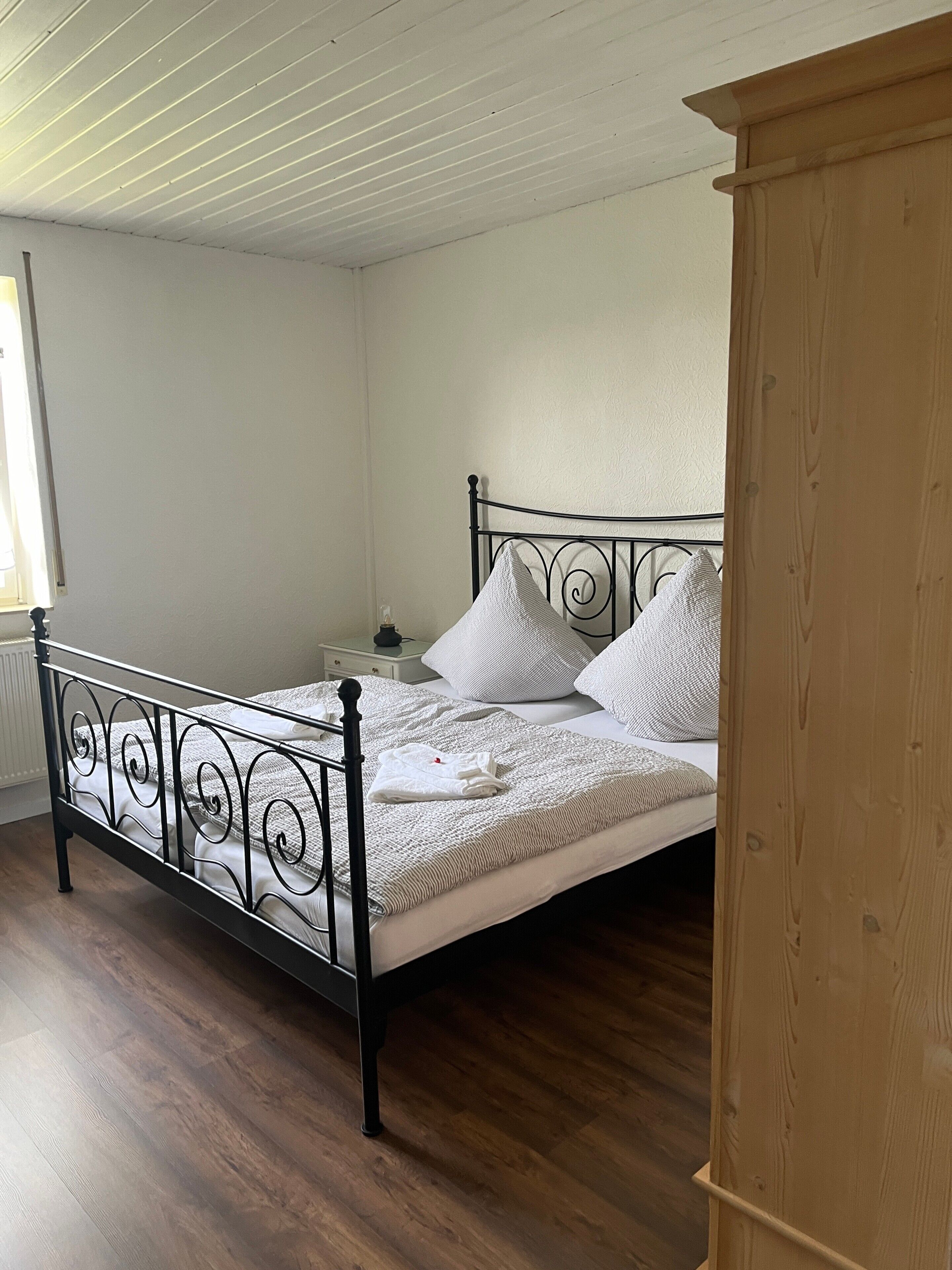 A single bed with a metal frame, a wooden nightstand, and a wooden wardrobe in a room with white walls and wooden flooring.