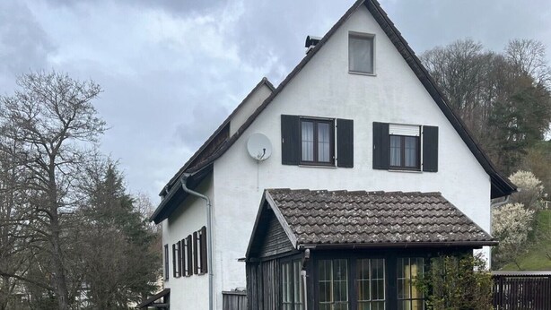 Une maison blanche avec une fenêtre à cadre noir, un petit jardin avec des fleurs violettes et un étroit canal d’eau à l’avant.