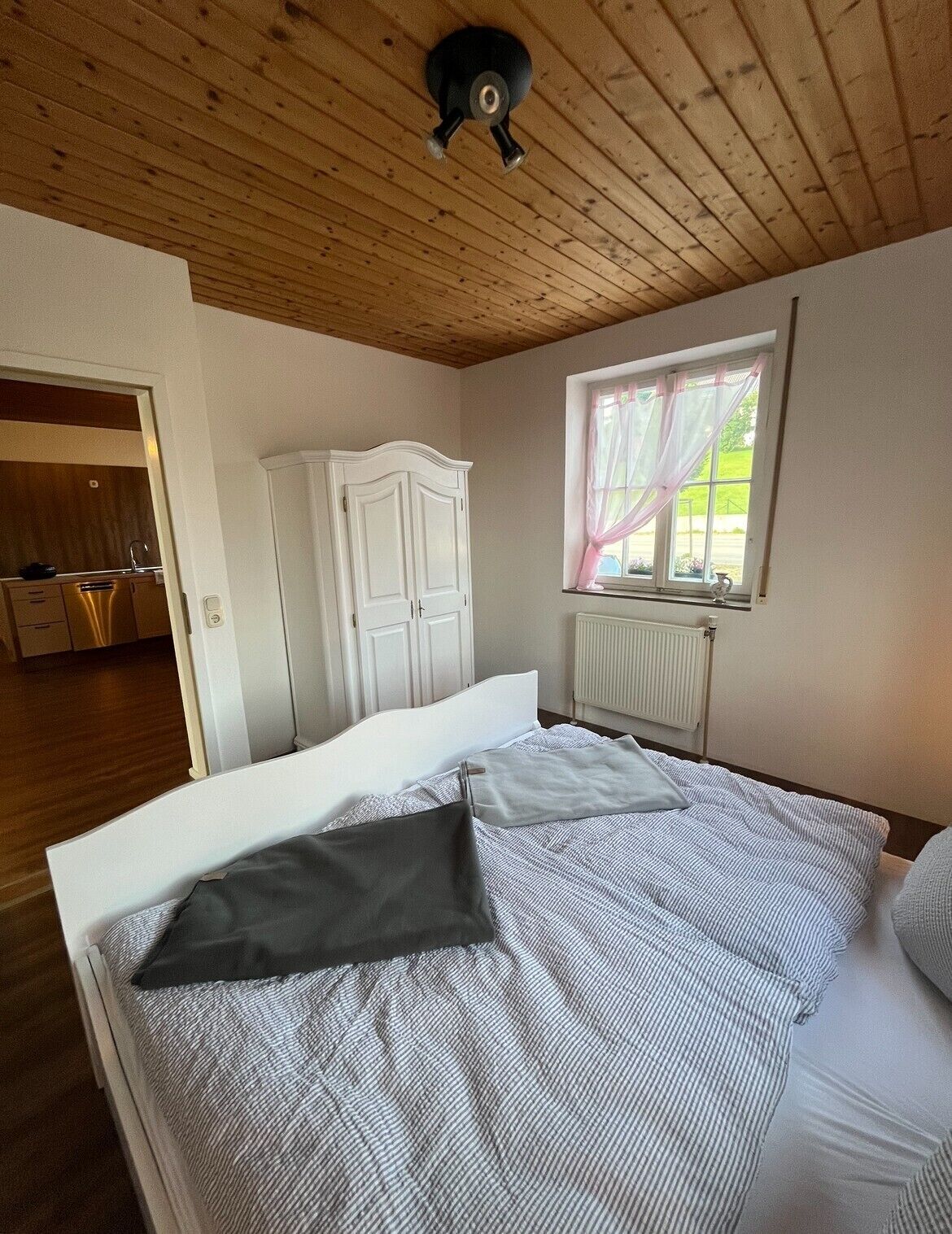 Une chambre avec un plafond en bois, un lit avec une literie à rayures, une armoire blanche et une fenêtre avec des rideaux.