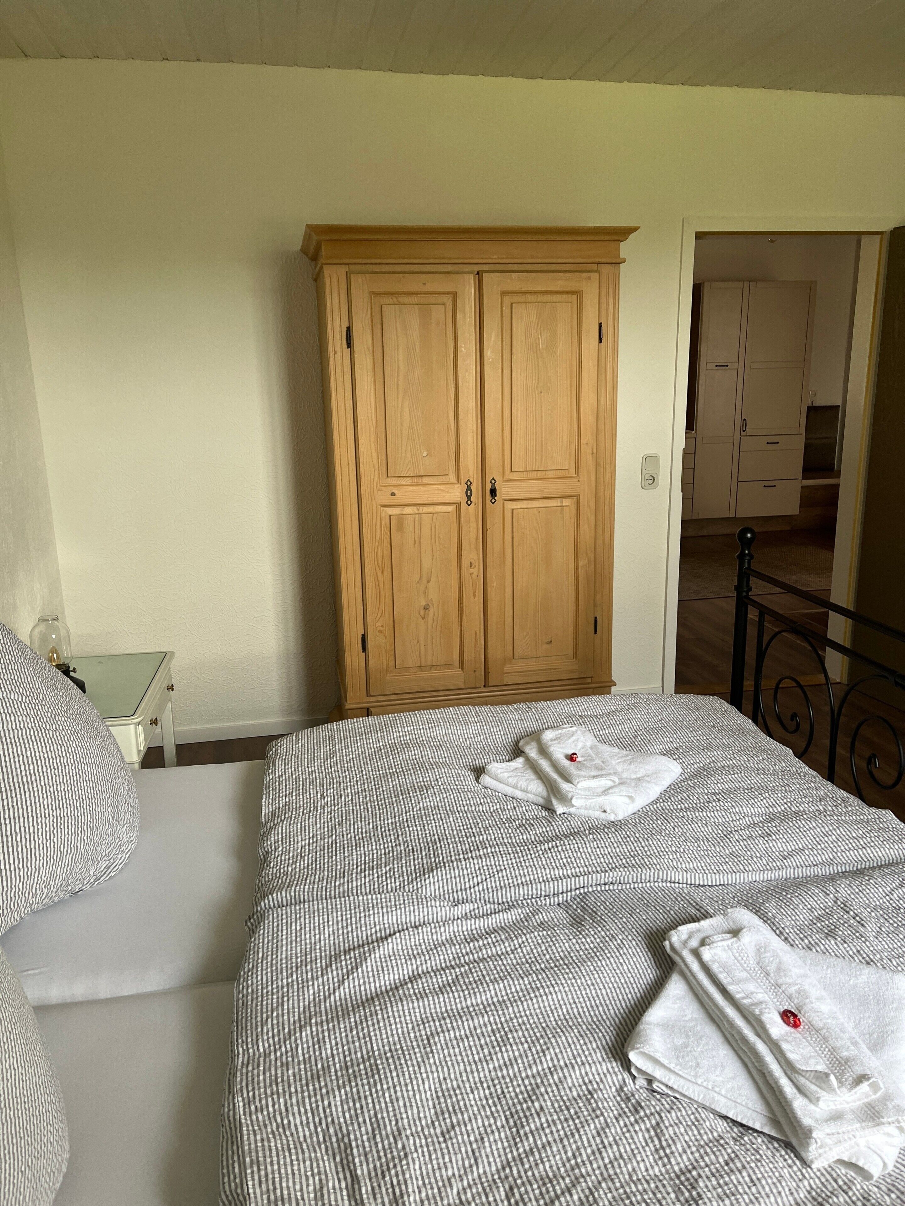A bedroom with a bed, pillows, a wooden wardrobe, and a small table.
