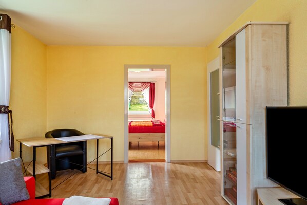 Una habitación con escritorio, silla, televisor, armario y cama con colcha roja.