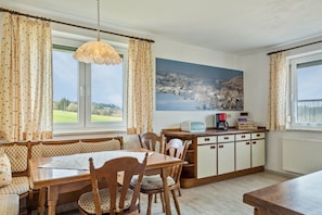 Un espace repas comprenant une table et des chaises en bois, un banc, un comptoir de cuisine avec des appareils électroménagers et une grande fenêtre avec des rideaux.