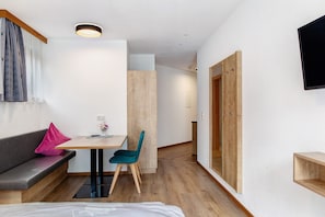 A modern room with a wooden floor, a desk with a chair, a sofa with a cushion, a wall-mounted TV, and a wooden wardrobe.