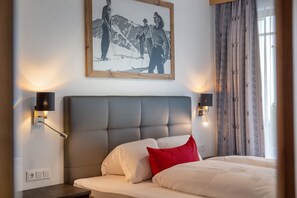 Une chambre d’hôtel avec un grand lit, une tête de lit, deux lampes fixées au mur, un tableau encadré accroché au mur et une fenêtre avec des rideaux.