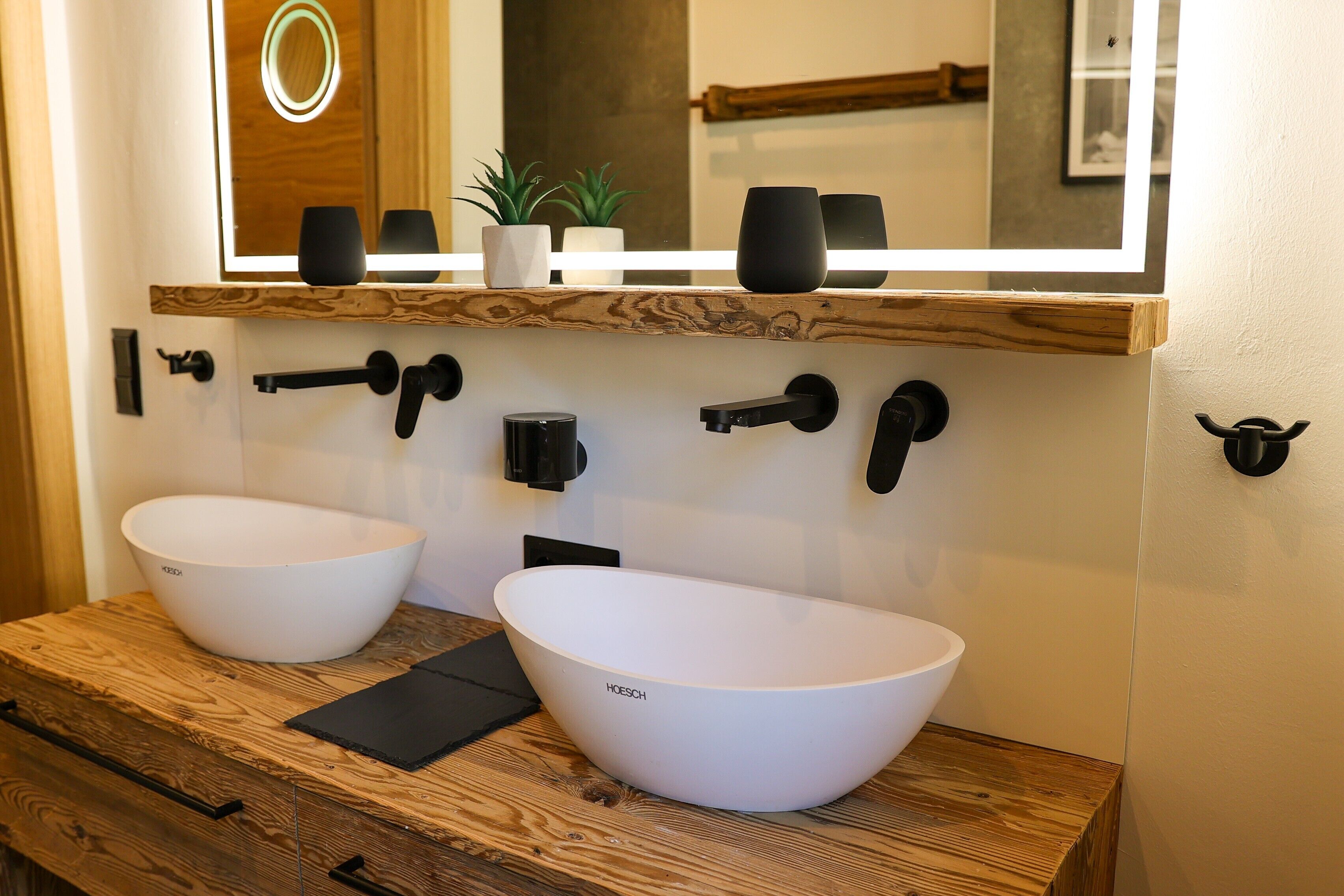 A bathroom with two white sinks, a wooden vanity, a mirror with a light, and a small potted plant.