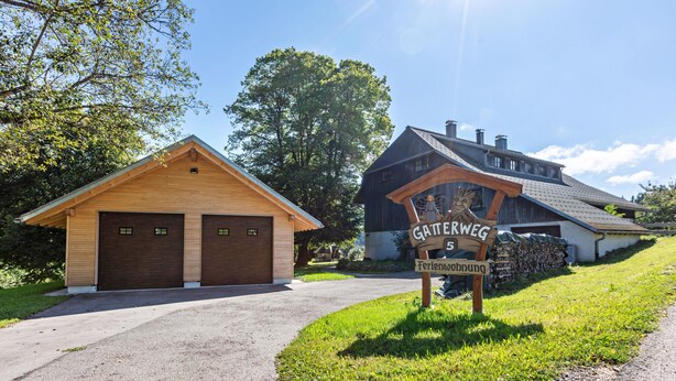 Une cabane en bois avec deux portes de garage, un panneau indicateur portant les inscriptions « Gatterweg » et « Ferienwohnung », et un bâtiment plus grand en arrière-plan.