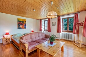 A room with a wooden ceiling, a bed, a sofa, a table with a plant, a radiator, and a window with curtains.