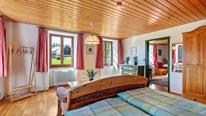 Une chambre avec un plafond en bois, un lit recouvert d’une courtepointe, une commode en bois, un radiateur et une fenêtre avec des rideaux.