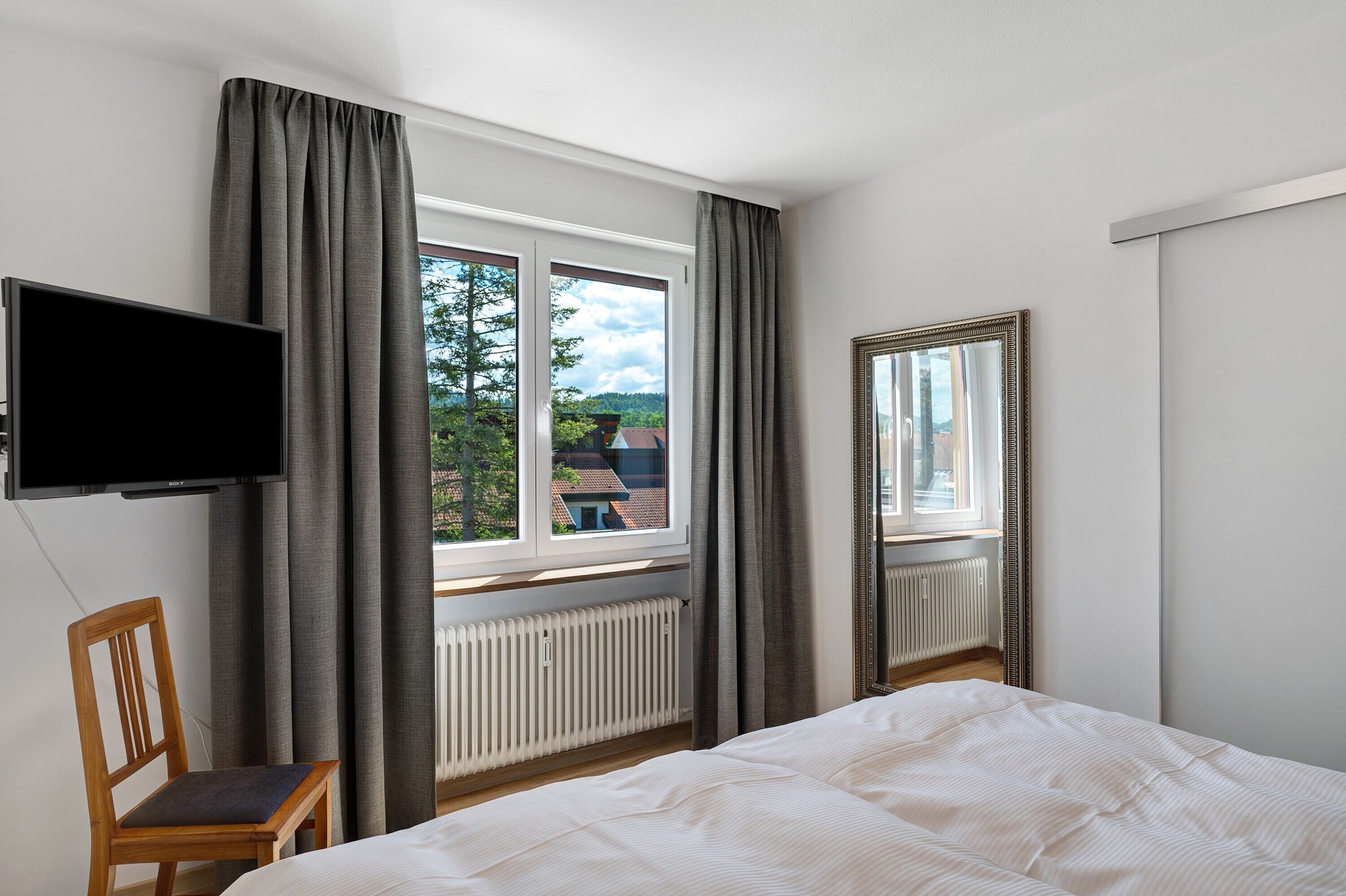 Een slaapkamer met een bed, een stoel, een televisie en een raam met gordijnen.