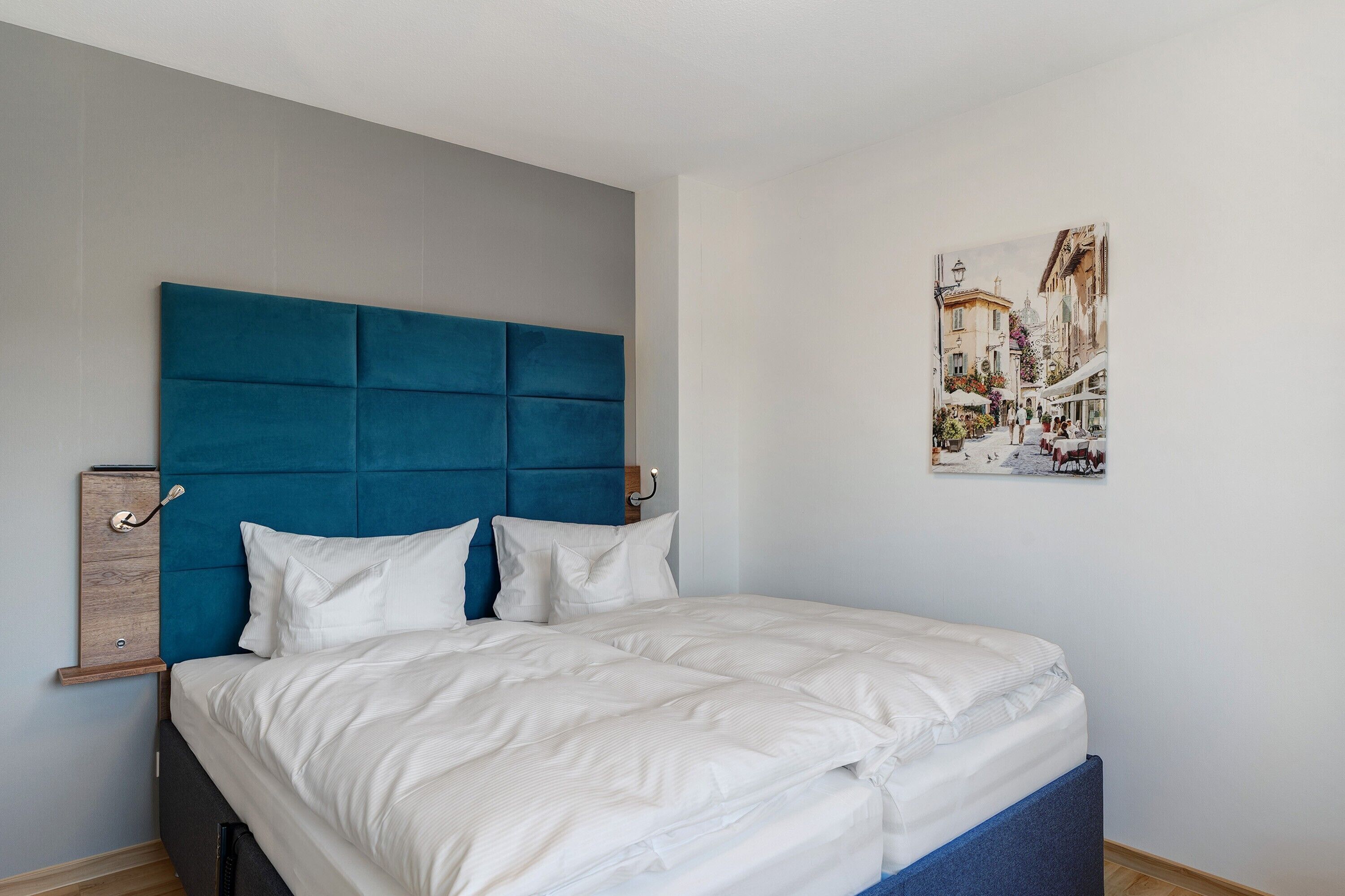 A bedroom with a blue headboard, white bedding, and a cityscape painting on the wall.