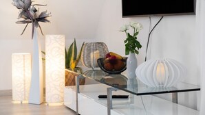 Uma sala de estar moderna com uma mesa de centro de vidro, um vaso branco com flores, um abajur decorativo e uma TV de tela plana na parede.