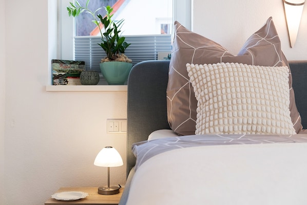 A neatly made bed with a textured pillow, a bedside lamp, a small potted plant, and a framed picture on the nightstand.
