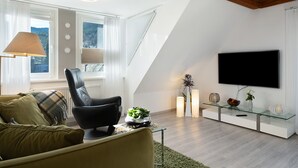 Uma sala de estar moderna com um sofá, uma poltrona, uma mesa de centro de vidro, uma TV e uma janela com cortinas.
