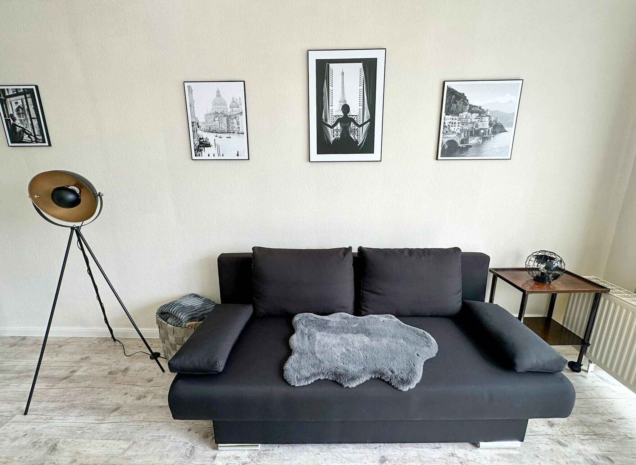 Een moderne woonkamer met een zwarte bank, een vloerlamp en drie ingekaderde foto’s aan de muur.