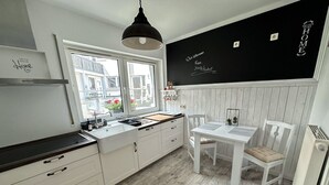 Een moderne keuken met witte kasten, een schoolbord waarop 'Welcome Home' staat, een raam met uitzicht op gebouwen, en een eethoek met een witte tafel en stoelen.
