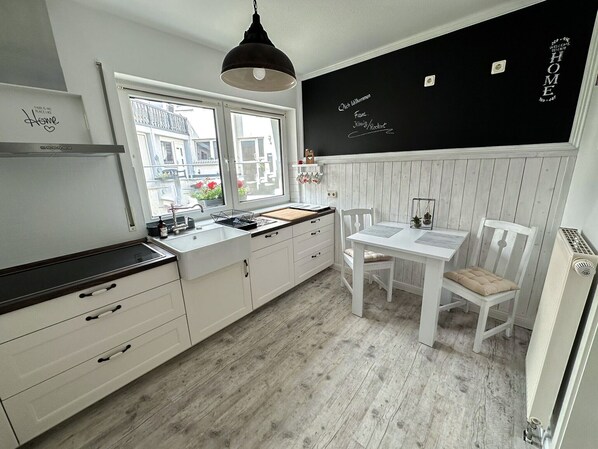 A modern kitchen with white cabinetry, a blackboard with 'Welcome Home' written on it, a window with a view of buildings, and a dining area with a white table and chairs.