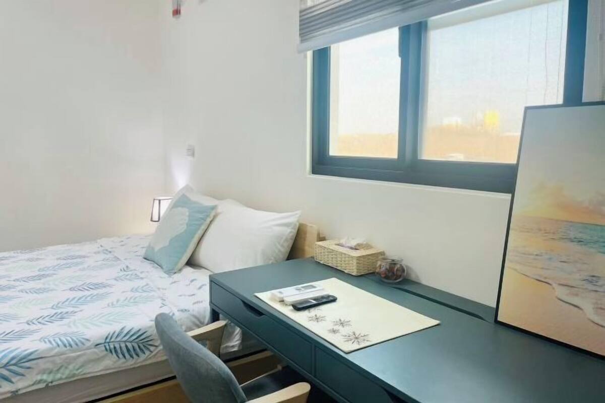 A bedroom with a bed, a desk, a chair, a window, and a painting.