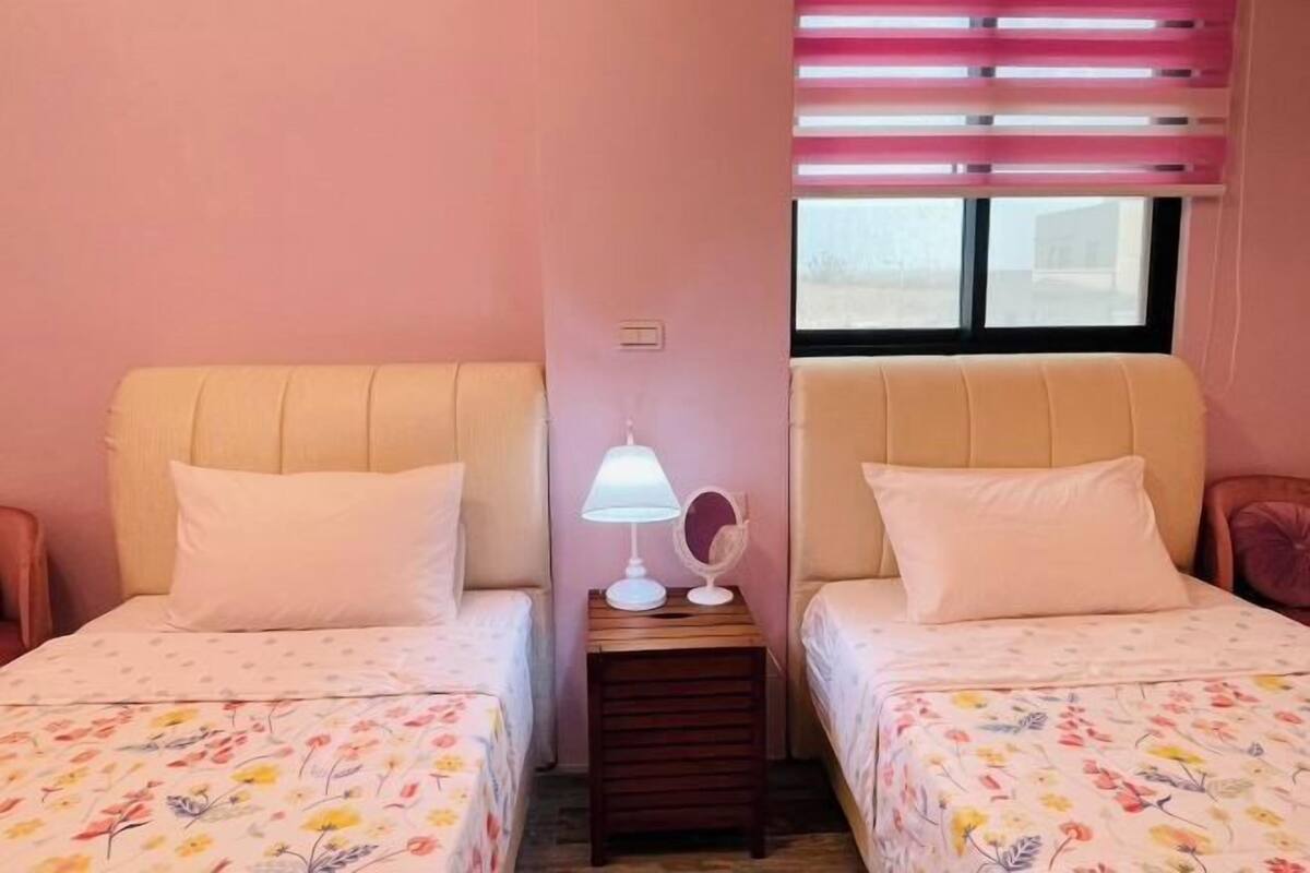 A room with two beds, a nightstand with a lamp, a mirror, and a window with blinds.