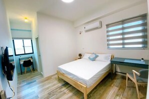 Un dormitorio con cama, escritorio y silla. Hay una ventana con persianas, un equipo de aire acondicionado y un televisor en la pared.