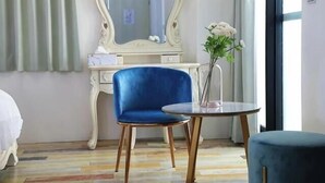 A hotel room with a blue chair, a round table, a white dresser with a mirror, and a vase with flowers.