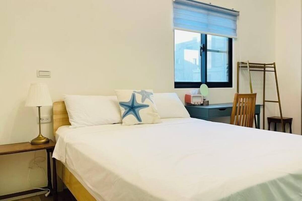 A hotel room with a bed, a bedside table with a lamp, a desk with a chair, a window with blinds, and a wooden chair.