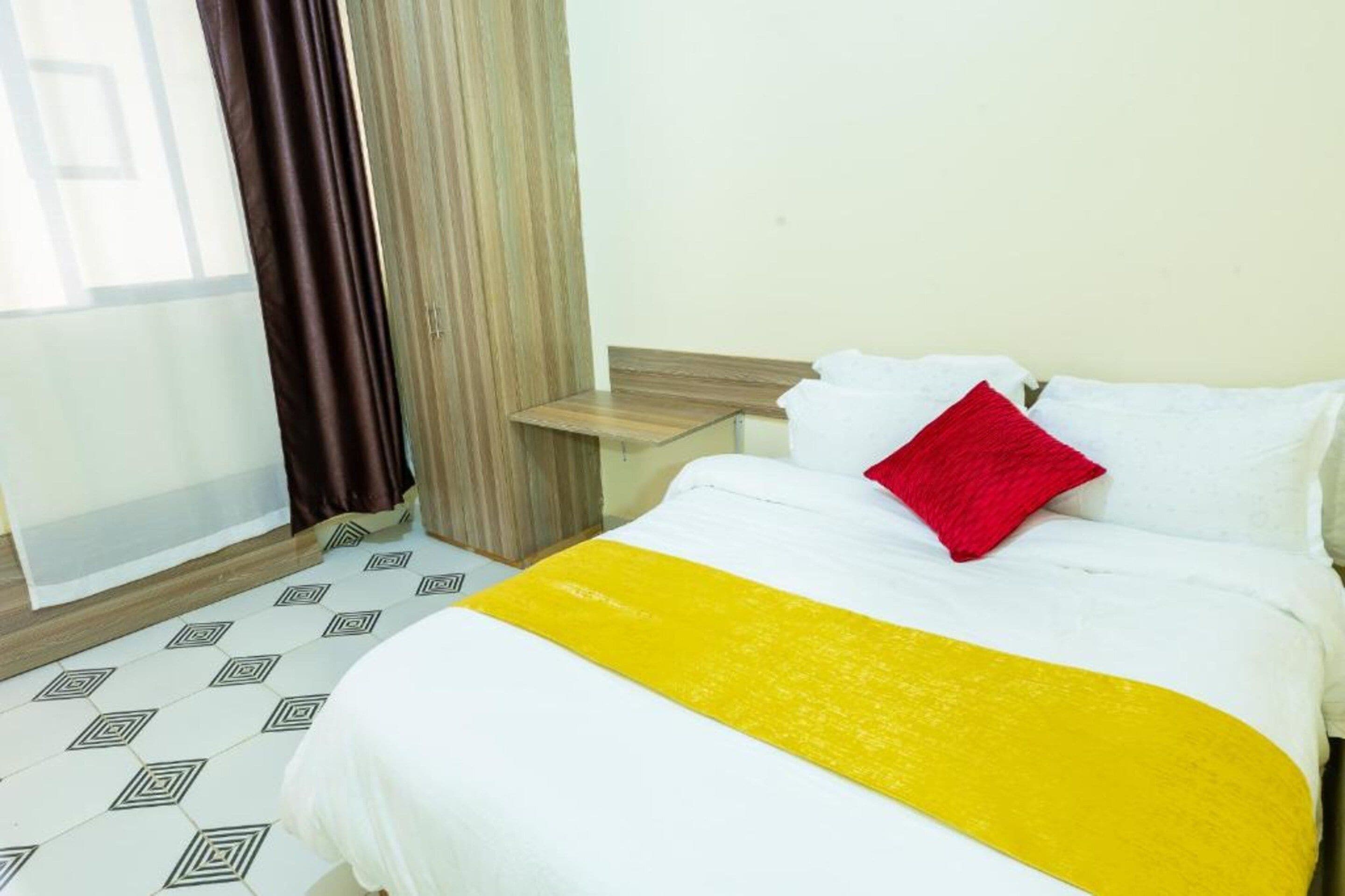Une chambre d’hôtel avec un lit, une table de chevet en bois, une fenêtre avec des rideaux et des carreaux de sol à motifs.