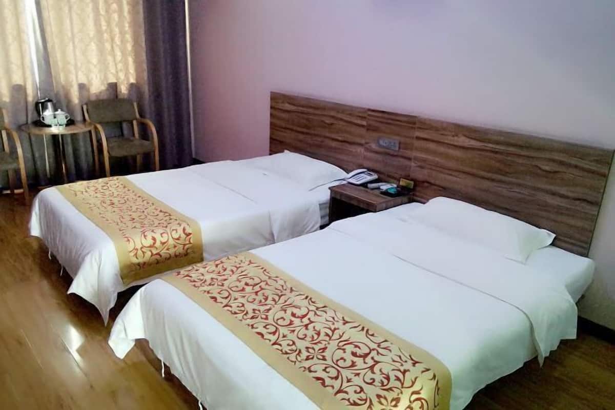 A hotel room with two beds, a wooden headboard, a small table with a kettle, and a chair.