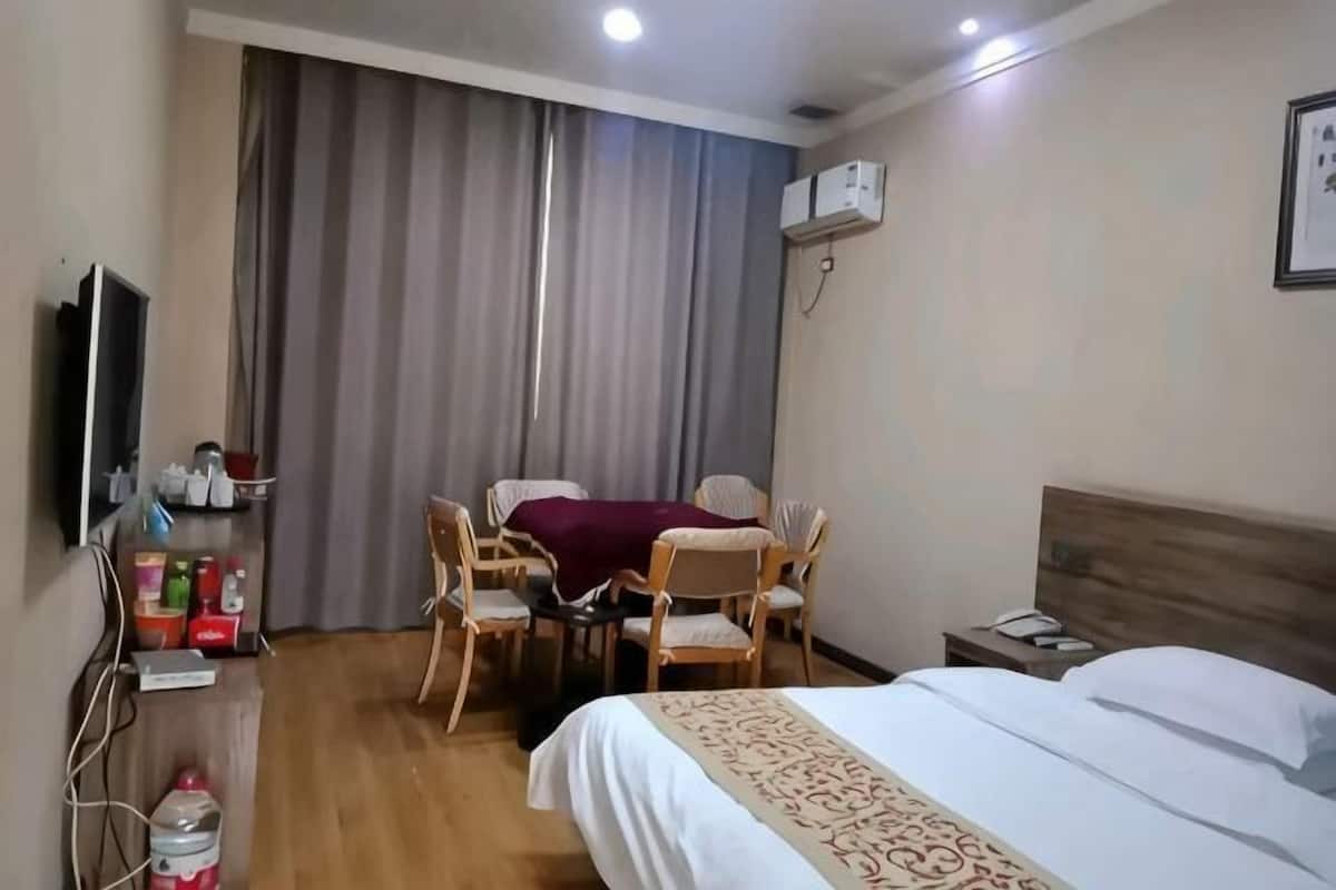 A hotel room with a bed, a desk with a chair, a table with a chair, a TV, and a window with curtains.