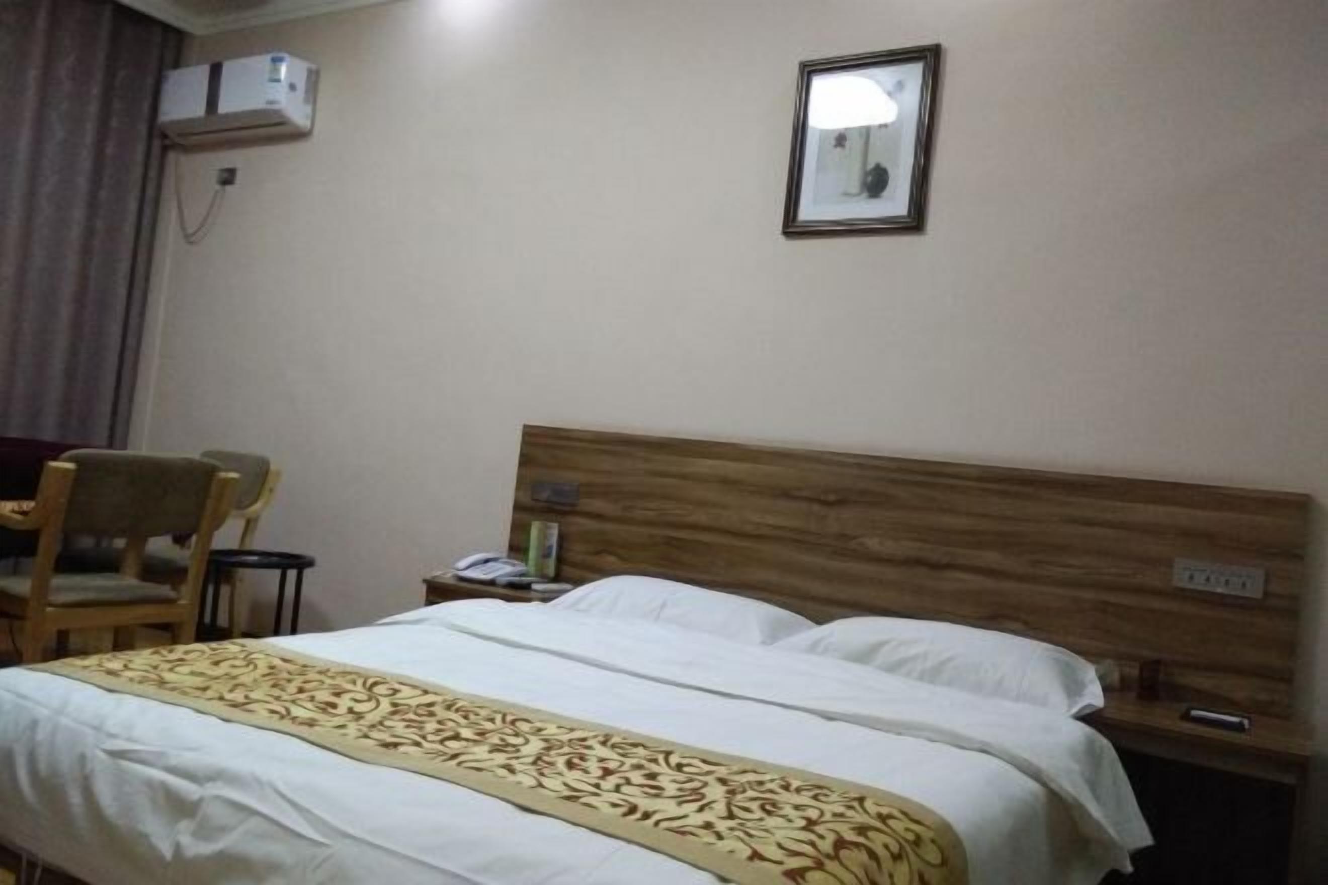 Una habitación de hotel con cabecera de madera, cama con sábanas blancas y un plaid estampado, una silla, una mesita de noche y un equipo de aire acondicionado en la pared.