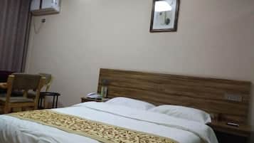 Una habitación de hotel con cabecera de madera, cama con sábanas blancas y un plaid estampado, una silla, una mesita de noche y un equipo de aire acondicionado en la pared.