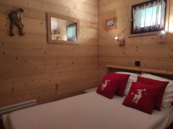 Een houten slaapkamer met een bed, rode kussens, een spiegel en een raam met gordijnen.