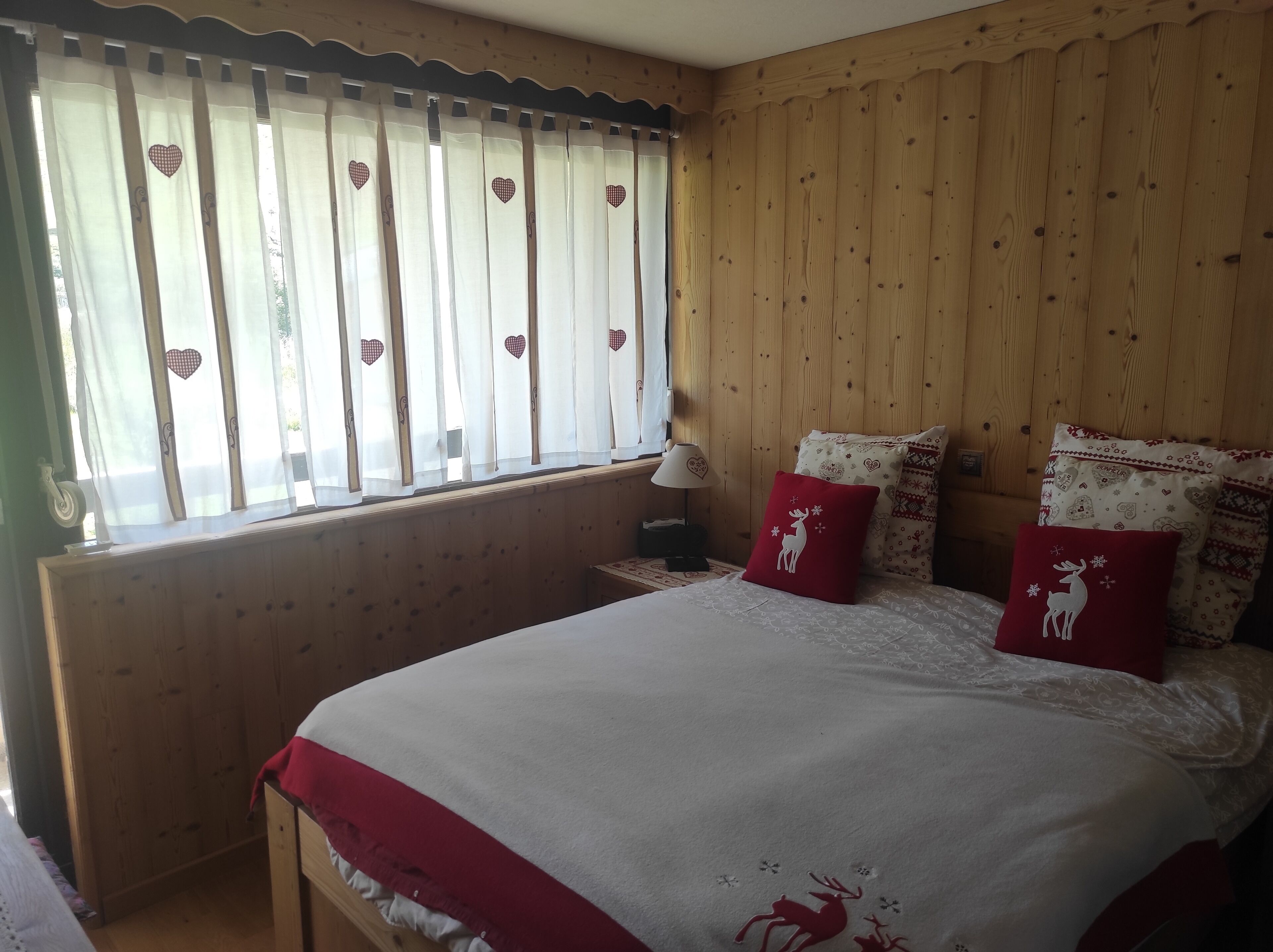 Een knus slaapkamer met een houten lambrisering, een bed met wit en rode beddengoed, en gordijnen met een hartpatroon.