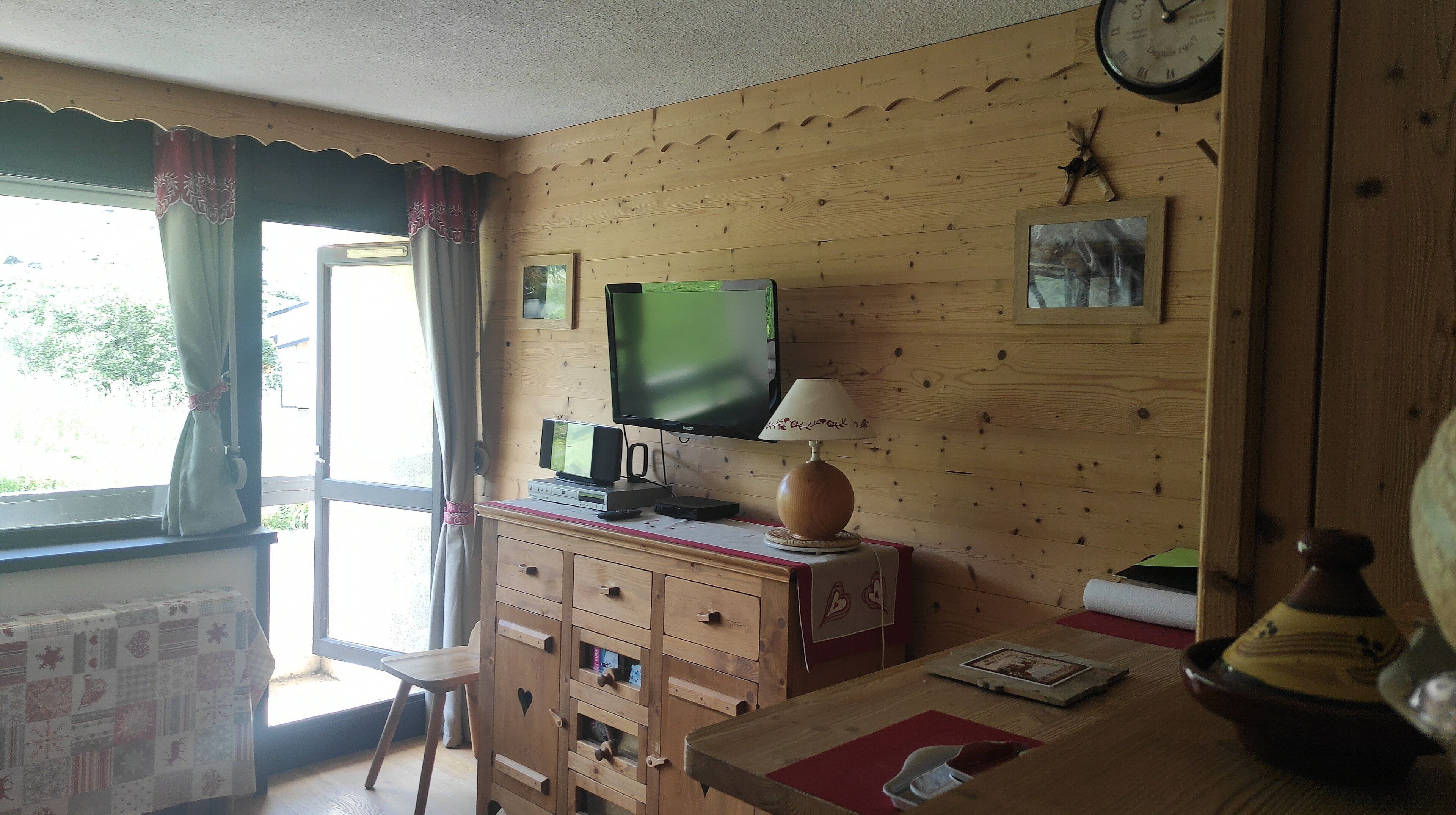 Una habitación acogedora con paredes de madera, un televisor, una lámpara, un reloj y una ventana con cortinas.