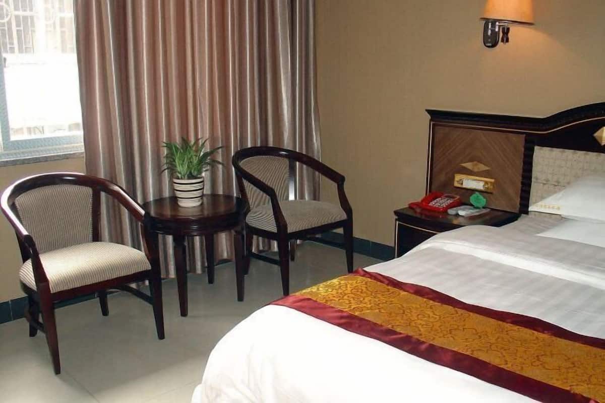 Een hotelkamer met een bed, twee stoelen, een kleintje tafeltje met een plant, een lamp en een nachtkastje met een telefoon en een rood voorwerp.