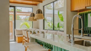 A modern kitchen with a long island, bar stools, a built-in sink, and large windows with greenery outside.