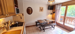 A kitchen with wooden cabinets, a microwave, a sink, and a dining table set for two with a bench. - Charming, well-equipped 4/6 pers Apt (Les Deux Alpes)