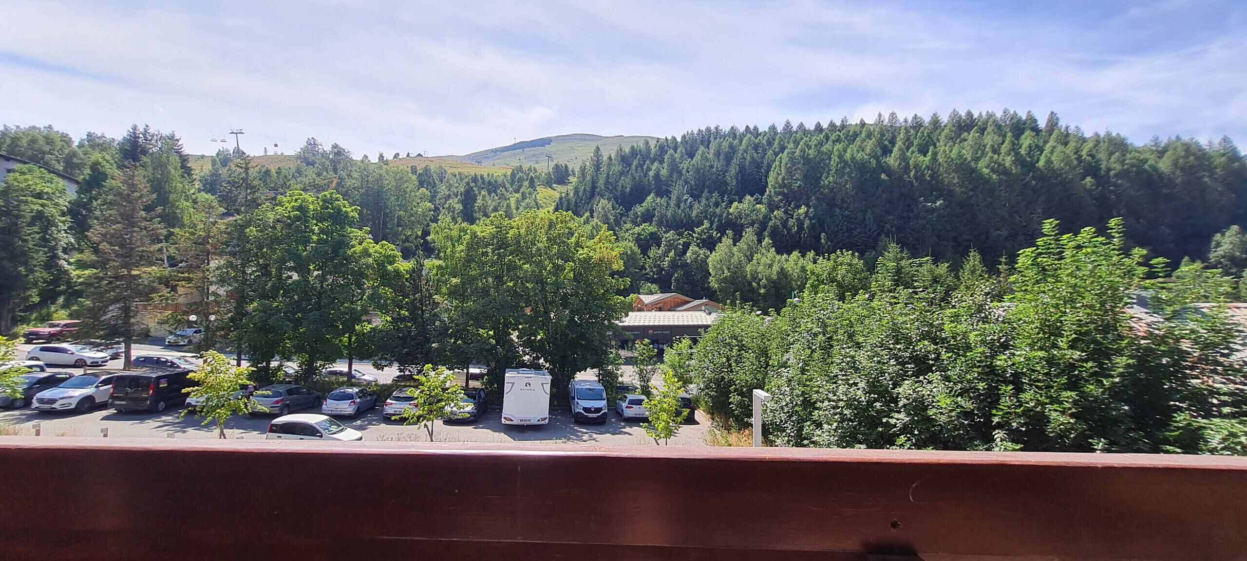 Un parcheggio con diverse auto e un camper bianco. Si vedono alberi e una collina con fitta vegetazione sullo sfondo.