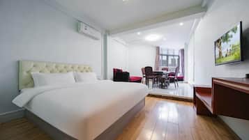 A hotel room with a large bed, a dining area with chairs and a table, a TV mounted on the wall, and an air conditioning unit.
