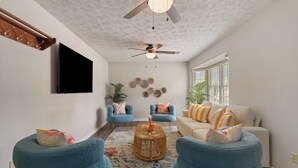 Una sala de estar con ventilador de techo, televisor de pantalla plana, dos sillones azules, un sofá beige y una mesa de centro.