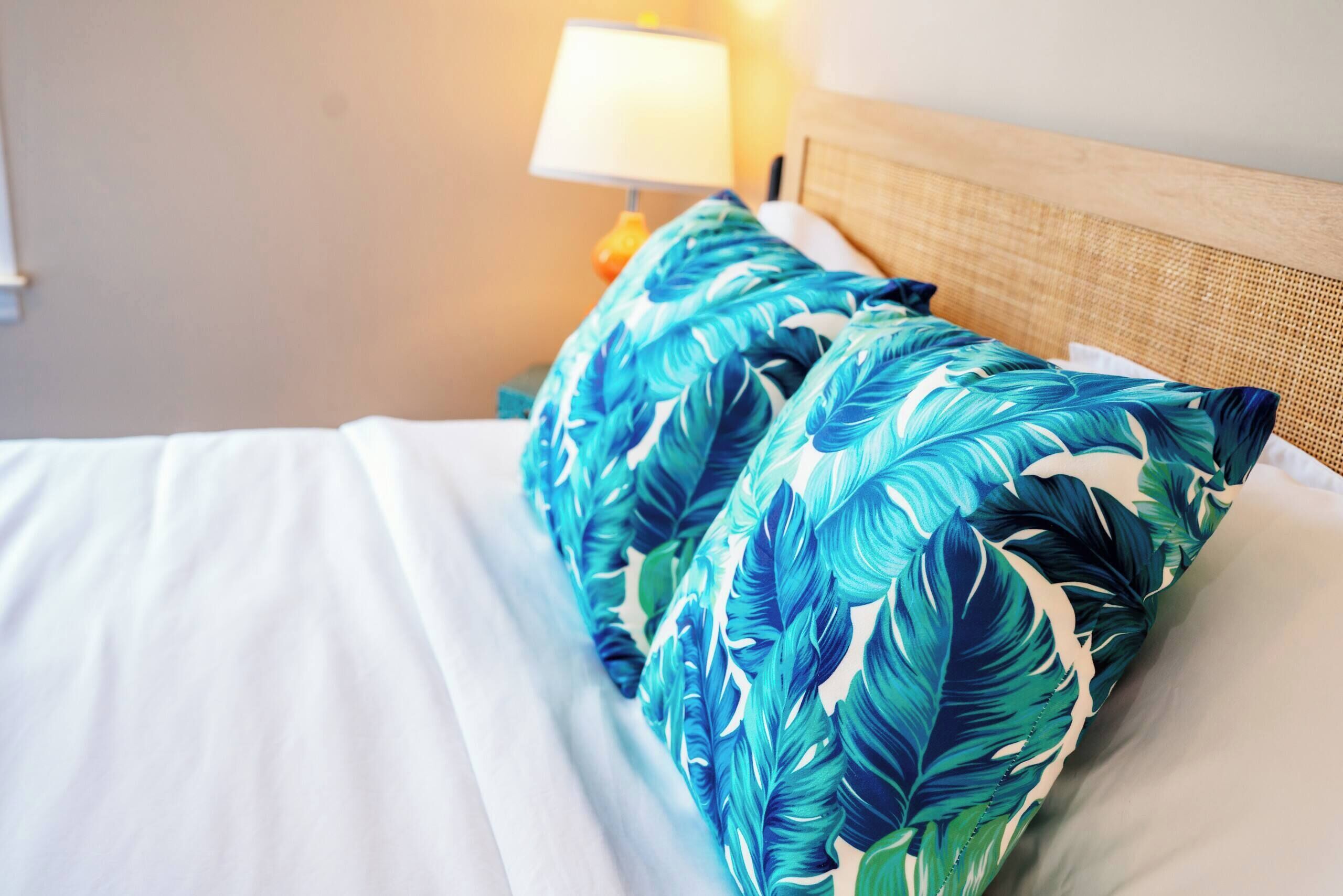 Una cama con ropa blanca de cama y un almohadón con diseño de hojas tropicales.