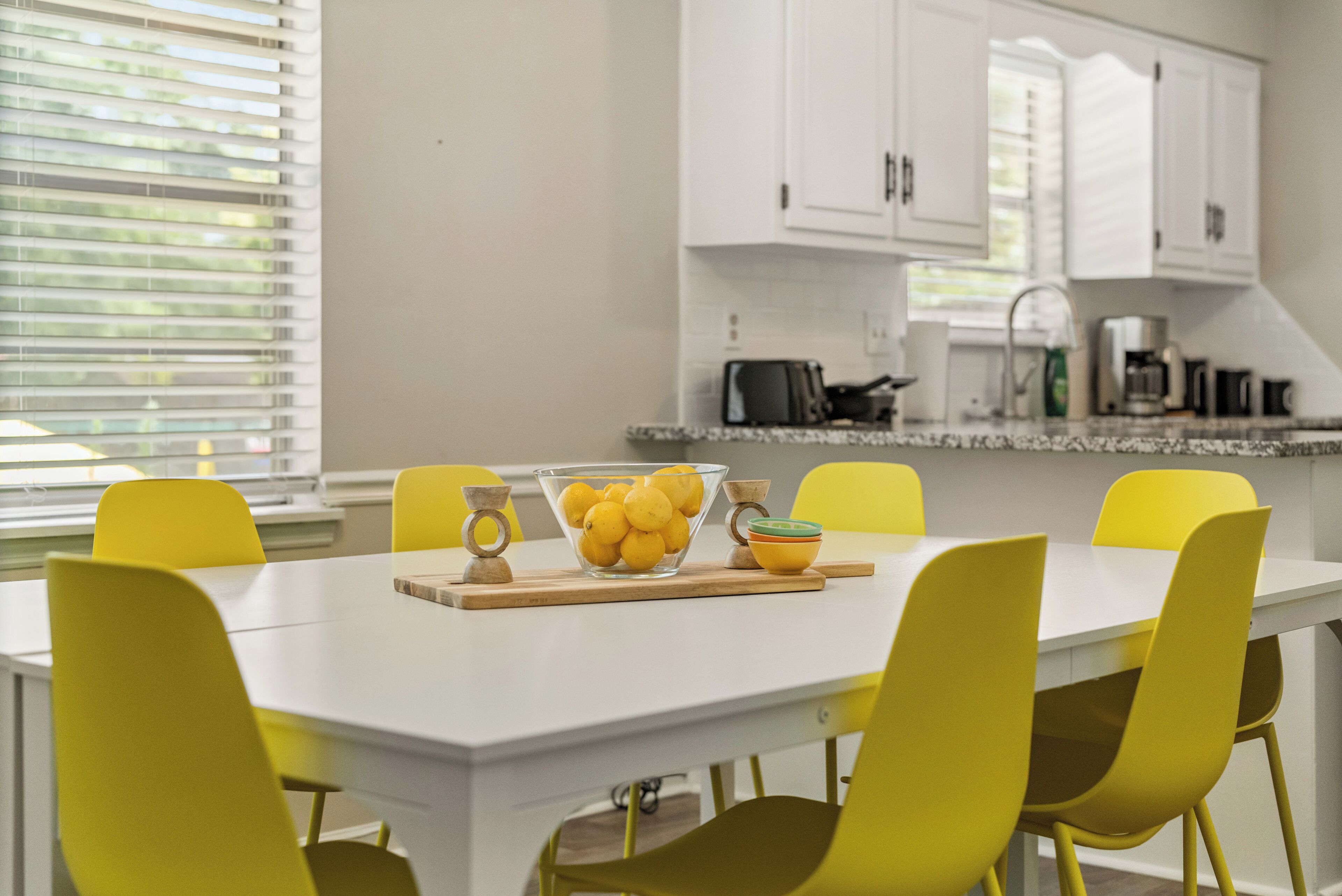 A bright kitchen with white cabinets, a white countertop, and yellow chairs around a central table with a bowl of lemons.