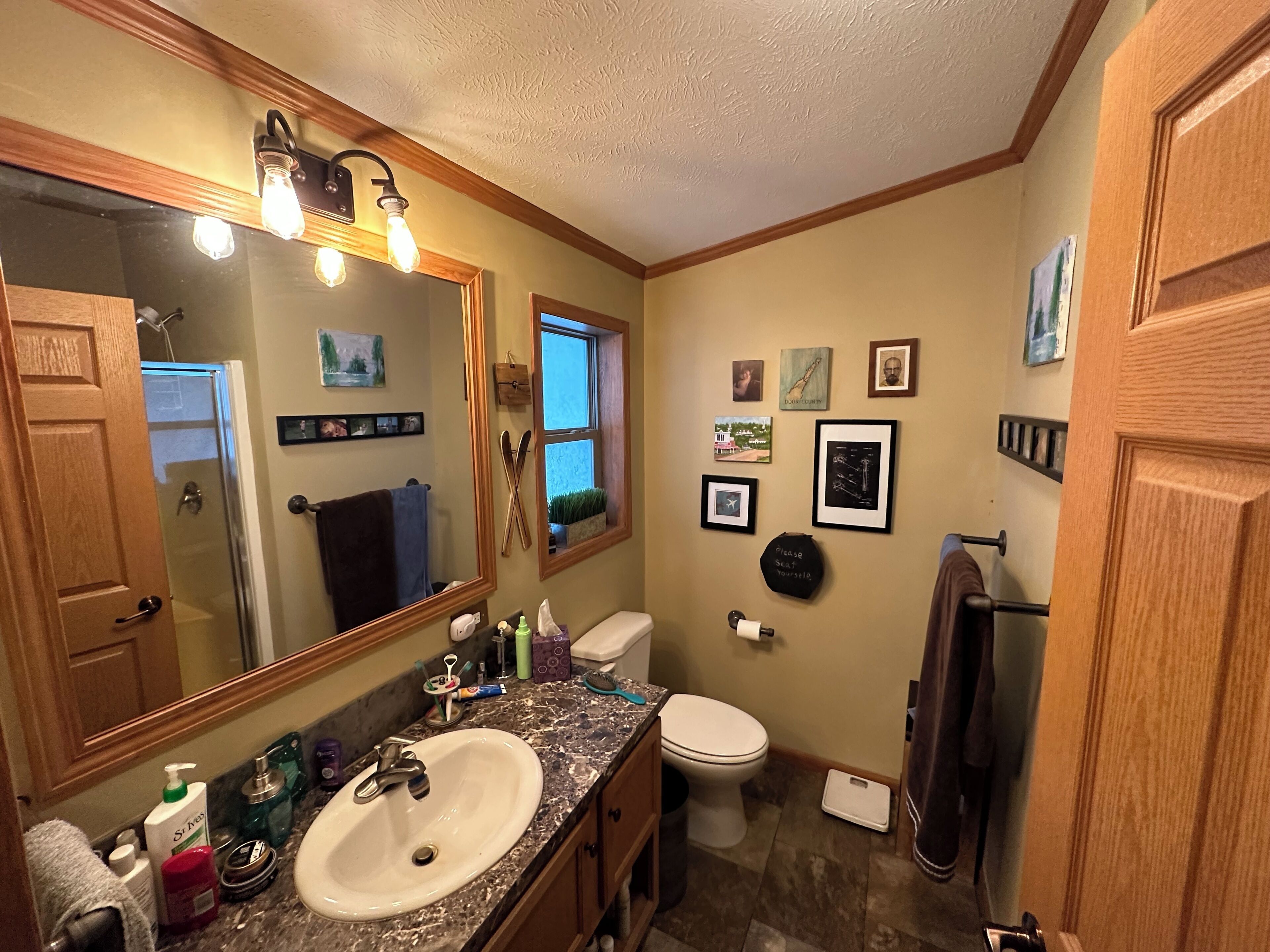 A bathroom with a sink, toilet, and mirror. There are framed pictures on the wall and a window with a view of the outside.