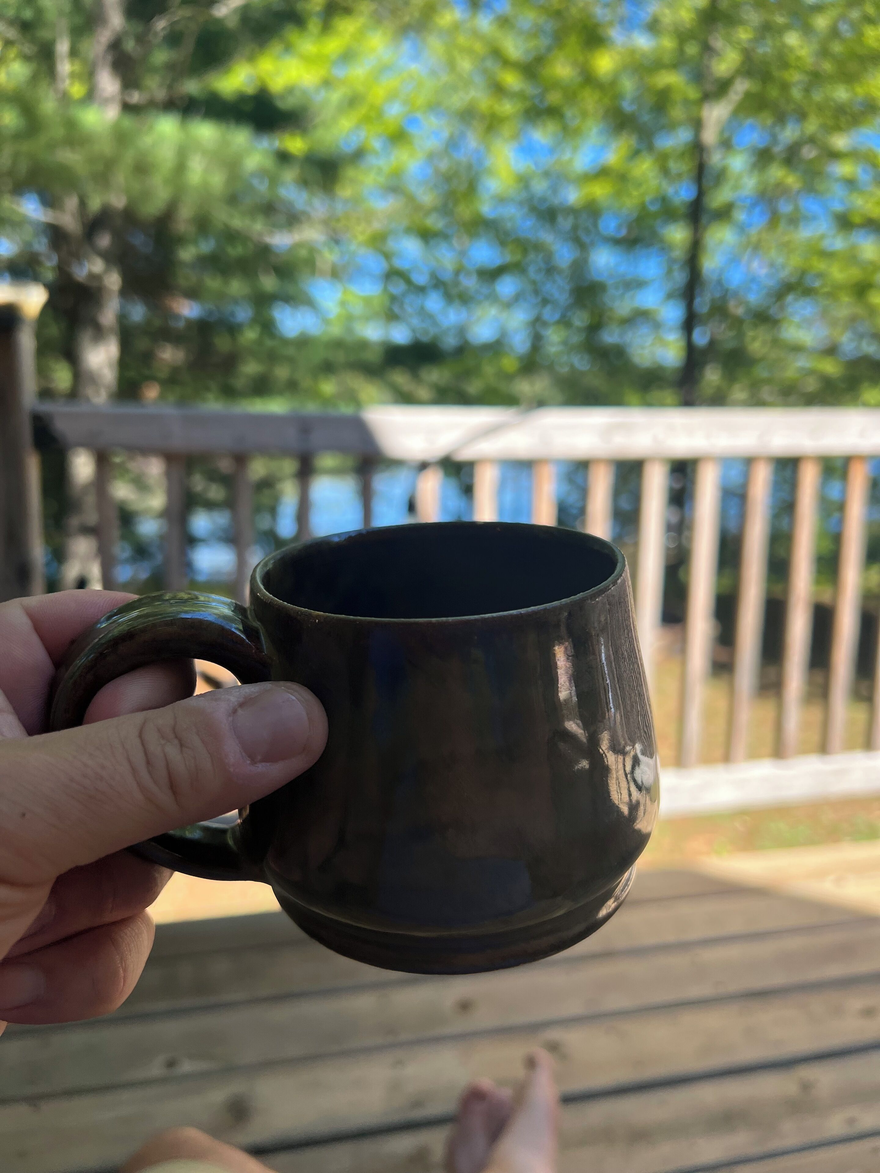 Eine Hand hält eine dunkel gefärbte Tasse auf einer Holzterrasse mit Geländer und Bäumen im Hintergrund.