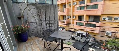 Un balcón con una mesa y sillas, una planta en maceta y vistas de un edificio de varios pisos con balcones.