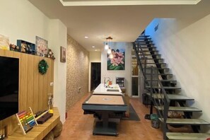 A living room with a TV, wooden entertainment unit, a pool table, a staircase, and a wall with a floral painting.