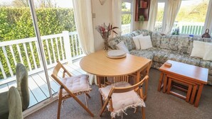 Una habitación acogedora con una mesa redonda de madera, dos sillas, un sofá y una pequeña mesita. Se ve un ventanal con cortinas y vistas al verdor.
